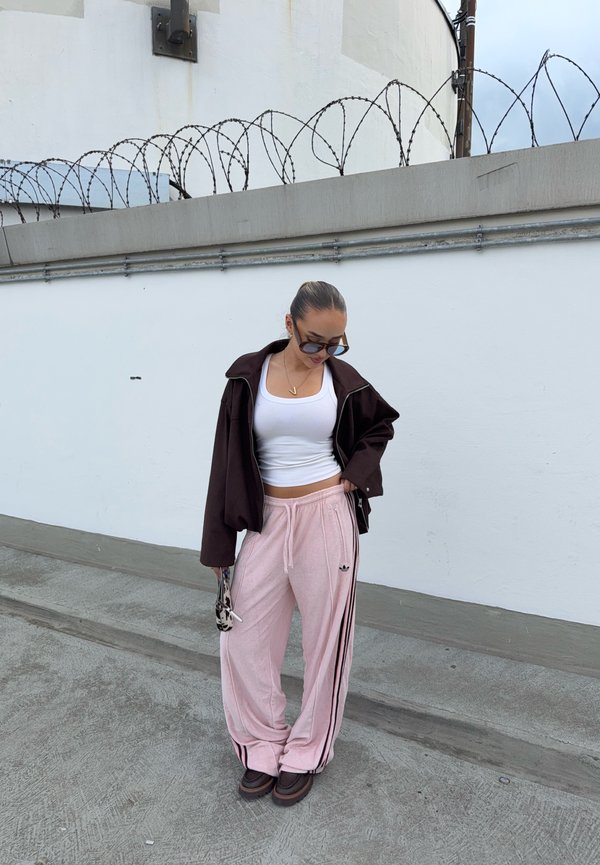 Mujer con gafas de sol, top corto blanco, chaqueta oscura y pantalones deportivos Adidas rosas, de pie en la acera cerca de una pared blanca coronada con alambre de púas.