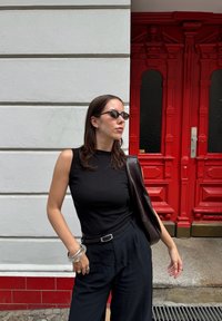 Svart ärmlös topp, mörka vida byxor och en brun läderjacka. Accessoarer inkluderar ett genomskinligt armband och solglasögon. Röd dörr i bakgrunden.