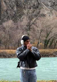 Veste aviateur en cuir noir, jean bleu clair, écouteurs blancs, debout près d'une rivière turquoise et d'arbres nus dans un paysage rocheux.