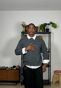 Maglione grigio lavorato a maglia con collo rotondo sopra una camicia bianca con colletto. Pantaloni neri. Gli accessori includono anelli e un braccialetto. Sfondo semplice.