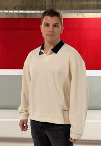 Bézs pulóver fekete gallérral, laza szabású, bordázott mandzsettákkal és egy kis fekete logó részlettel alul. Sötét farmerra van felvéve.