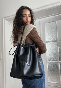 Sort læder bucket-tasker med prægede "CK"-logo, der har gyldne messingøjer og snørelukning. Modellen har en tætsiddende brun top på.