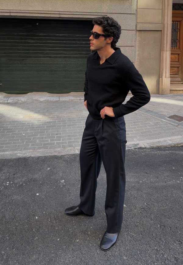 Mężczyzna w czarnym swetrze, szerokich czarnych spodniach, czarnych butach i ciemnych okularach przeciwsłonecznych stoi na miejskiej ulicy z rękami na biodrach.