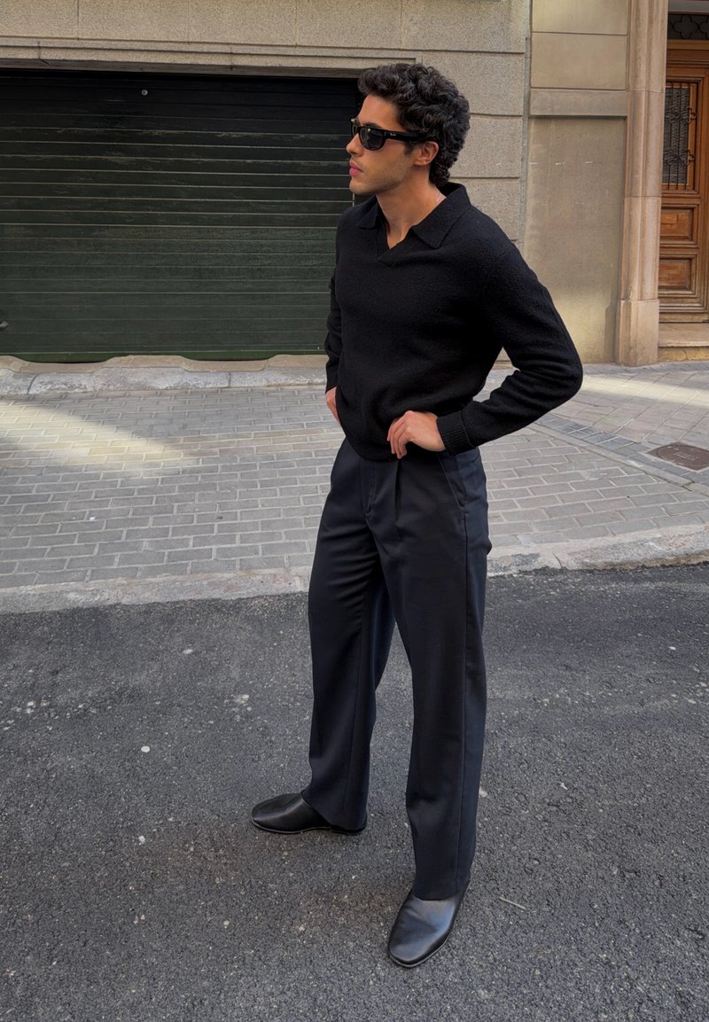 Homme portant un pull noir, un pantalon noir à jambes larges, des chaussures noires et des lunettes de soleil foncées, debout dans une rue urbaine les mains sur les hanches.