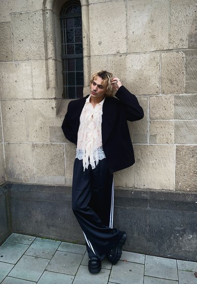 Blazer negro de gran tamaño, camisa de encaje con volantes en color crema, pantalones de satén negros con rayas blancas, zapatos negros gruesos, de pie contra una pared de piedra.