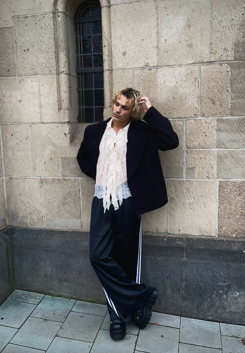 Μαύρο oversized σακάκι, κρεμ μπλούζα με δαντέλα και φρου φρου, μαύρο σατέν παντελόνι με λευκές ρίγες, χοντρές μαύρες γόβες, στέκοντας απέναντι σε μια πέτρινη τοιχοποιία.