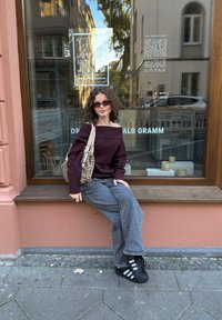 Une personne est assise sur un rebord en bois, portant un pull bordeaux à épaules dénudées, un jean gris à jambes larges et des baskets noires Adidas, tenant un sac à motifs.