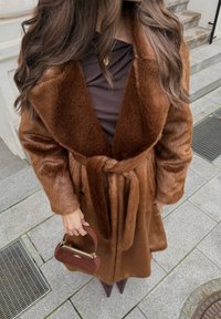 Brown faux fur coat with a wide collar, belted waist, and long sleeves. Brown top and a small brown handbag with a curved handle.