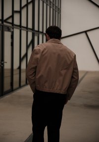Homme en veste beige et pantalon noir marchant à côté de fenêtres en verre sur une zone pavée extérieure avec des motifs architecturaux géométriques.