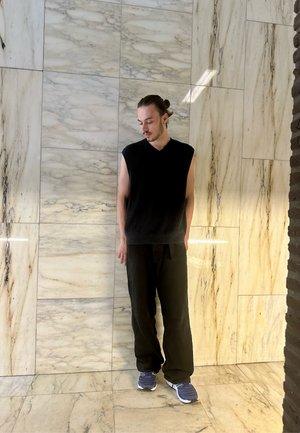 Suéter negro sin mangas, pantalones negros holgados y zapatillas deportivas azules. Frente a paredes de mármol claro texturizado.