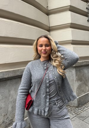 Joven mujer con largo pelo rubio que lleva un cárdigan gris y vaqueros, de pie junto a un edificio de piedra beige, con bolso rojo al hombro.