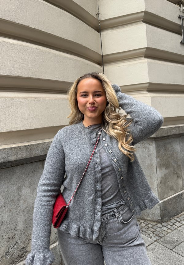 Młoda kobieta z długimi blond włosami, ubrana w szary kardigan i dżinsy, stojąca przy beżowym kamiennym budynku, czerwona torebka na ramieniu.