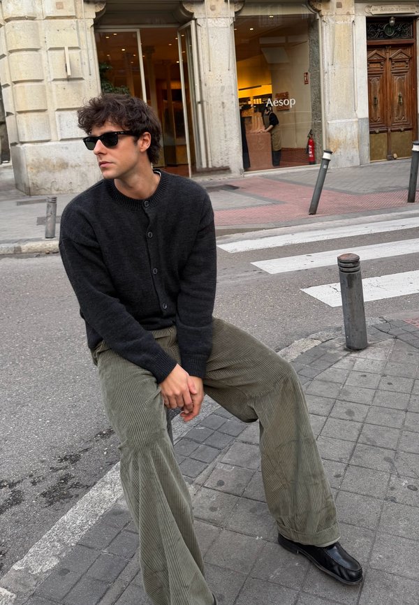 Črni pleteni kardigan čez sivo majico, kombiniran z širokimi, rebrastimi olivno zelenimi manšestrskimi hlačami in črnimi usnjenimi čevlji. Priložnostno ulično okolje.