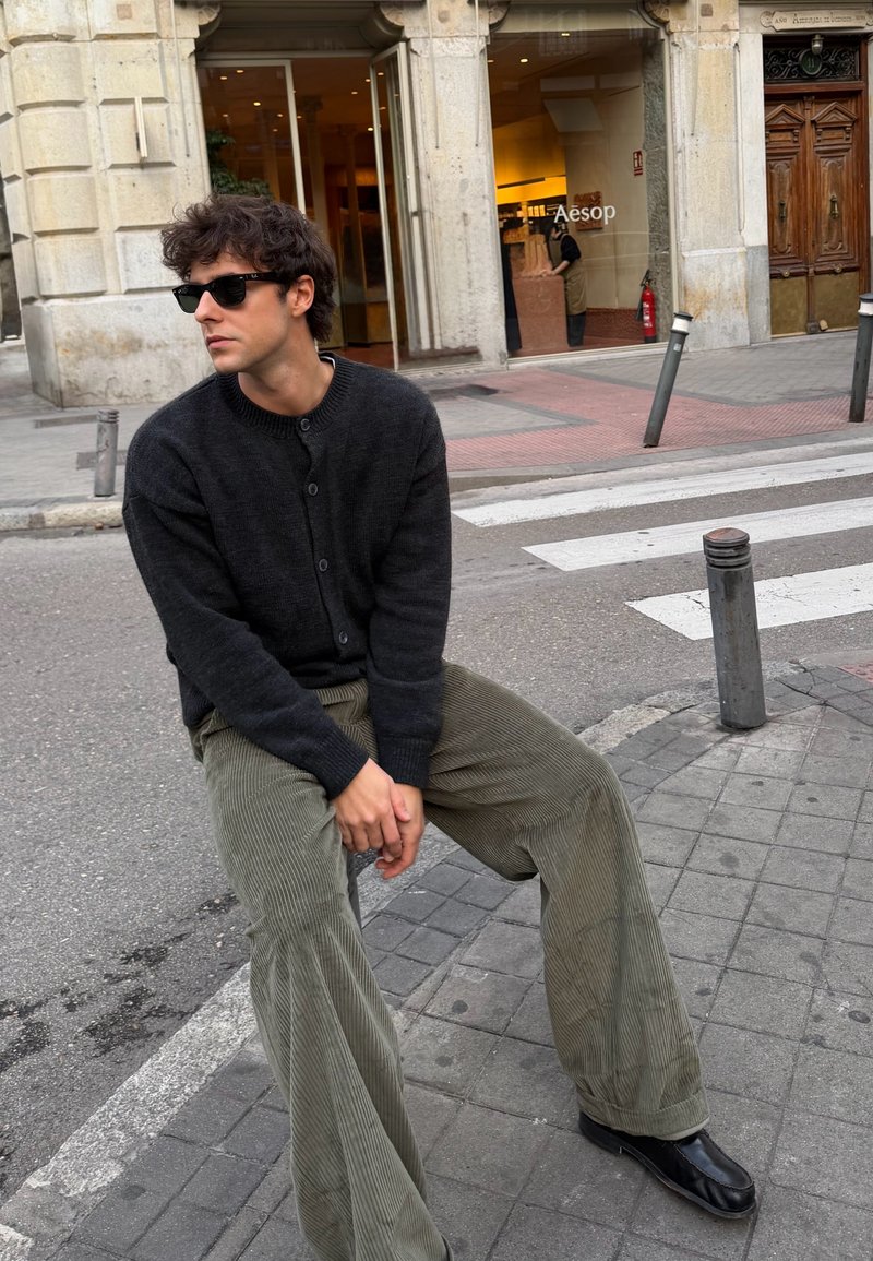 Zwarte gebreide cardigan over een grijze top, gecombineerd met wijde, geribde olijfgroene corduroy broek en zwarte leren schoenen. Informele straatsfeer.