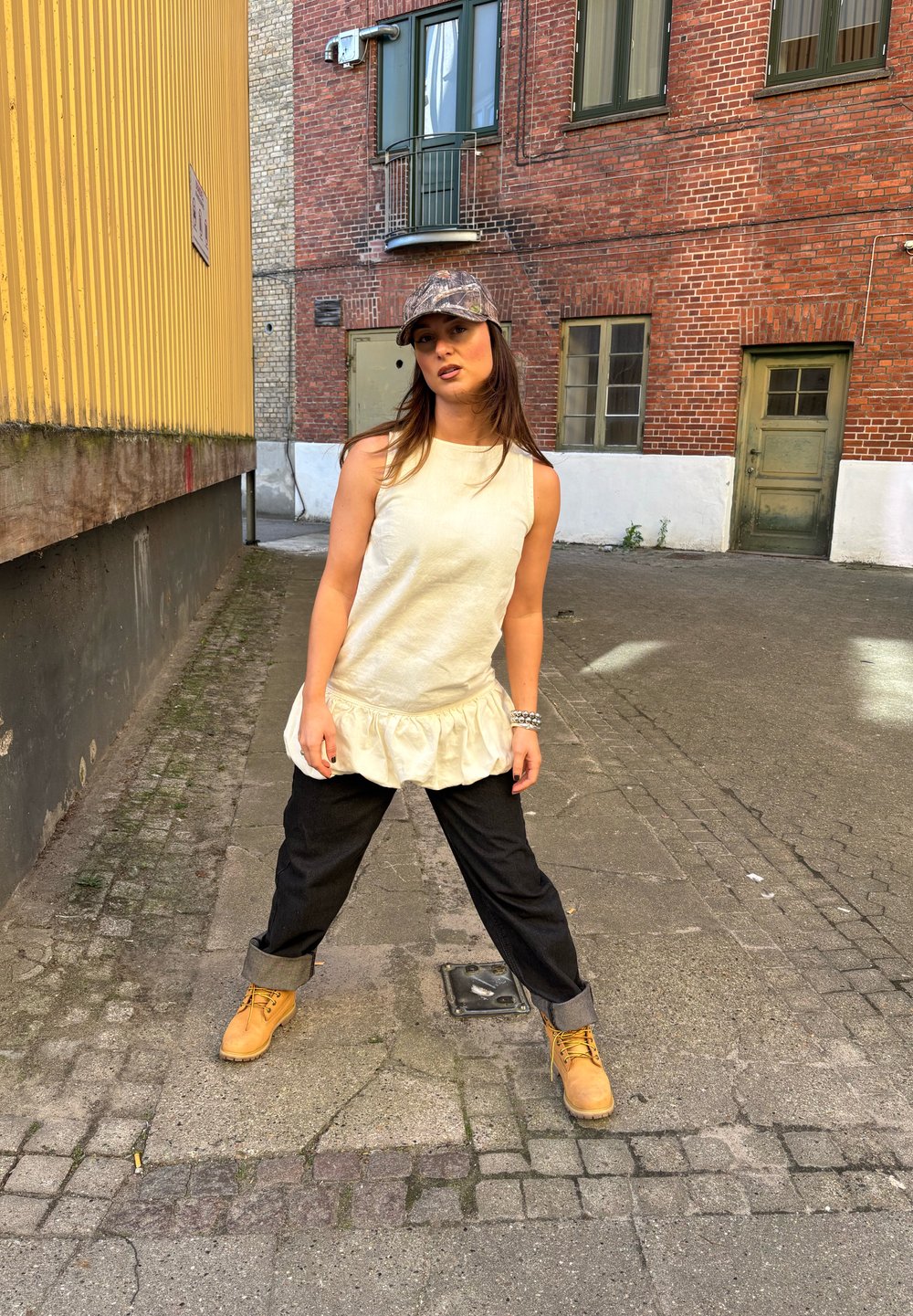 Beige ermeløs bluse med volangkant, kombinert med løse svarte jeans og brune støvler. Bakgrunnen har murvegger og et gult panel.
