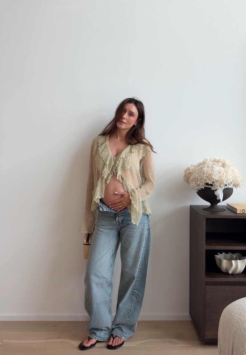 Femme enceinte en blouse transparente et jean, debout près d’un mur, la main sur le ventre, à côté d’un vase sombre avec des fleurs blanches sur une étagère en bois.