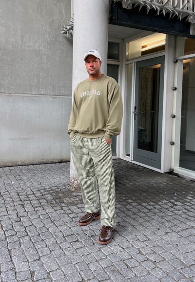 Sudadera verde oliva con el texto "EL PAPÁ", pantalones a rayas verdes y blancos, y zapatos de cuero marrón con suelas blancas. Fondo urbano.