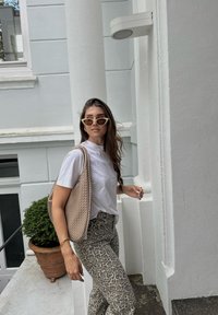 Světlý béžový tkaný tote batoh, bílá košile s krátkým rukávem a vzorované šedé legíny. Pozadí tvoří bílá zeď a květináč s rostlinou.