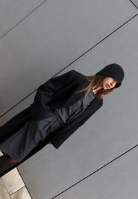 Femme portant un bonnet noir, un manteau noir, un haut gris et une jupe grise, debout contre un mur gris les mains dans les poches.