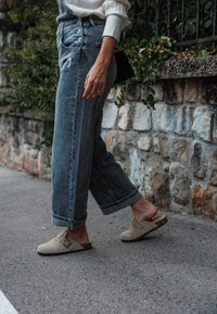 Jeans larges gris clair avec des déchirures, associés à des sabots beiges avec une boucle. En arrière-plan, un mur en pierre et de la végétation.
