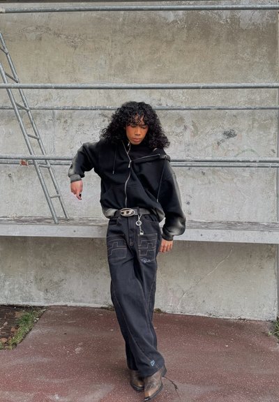 Sudadera negra de gran tamaño con cremallera, combinada con jeans anchos negros con costuras en contraste y bolsillos, y botas de cowboy marrones.