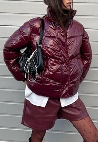 Femme portant une doudoune brillante bordeaux, un short en cuir assorti, des collants transparents à pois, une chemise blanche et un sac à main noir avec chaîne argentée.