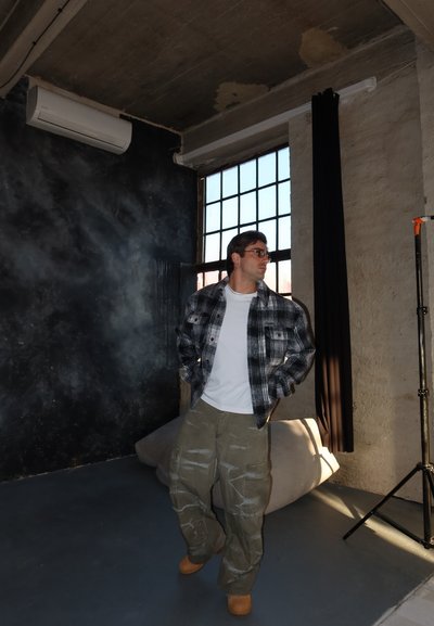 Hombre con chaqueta de cuadros, camisa blanca y pantalones cargo, de pie junto a una gran ventana en una habitación de estilo industrial con paredes expuestas.