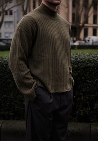 Pull en maille côtelée vert olive avec un col montant, caractérisé par une coupe ample et un ourlet brut, associé à un pantalon gris foncé dans un cadre décontracté.