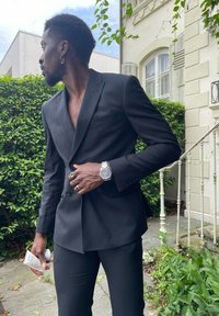 Zwarte op maat gemaakte blazer met een dubbelzijdig ontwerp, gecombineerd met bijpassende broeken. De persoon houdt een blik vast en draagt een zilveren horloge.