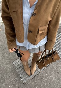 Brun suedeblazer med to knapper og lommer båret over en lyseblå skjorte; grå shorts; knæhøje beige støvler; holder sorte solbriller og en brun taske.