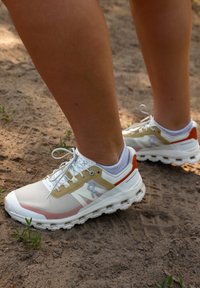 Witte en beige hardloopschoenen met een bovenwerk van mesh en oranje accenten. Beschikt over een gevoerde zool en platte veters voor ondersteuning.