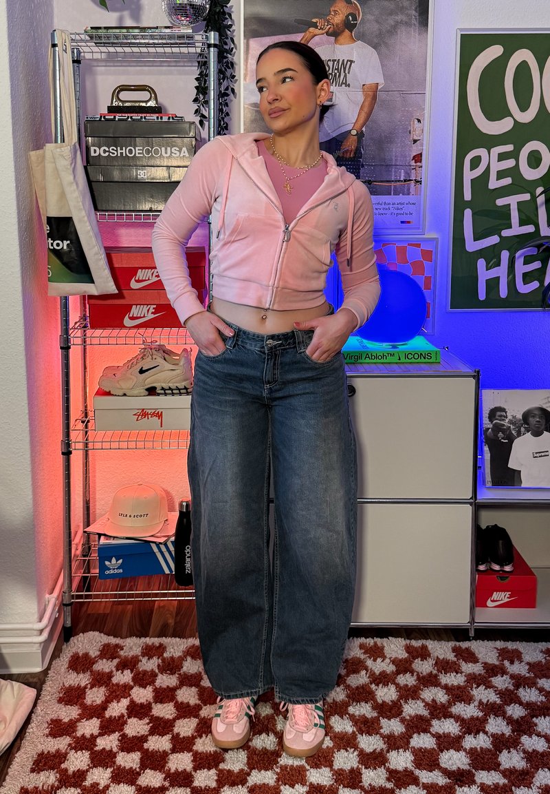 Donna con felpa corta rosa e jeans larghi, in piedi con le mani nelle tasche in una stanza con scatole di sneaker, poster e illuminazione colorata.