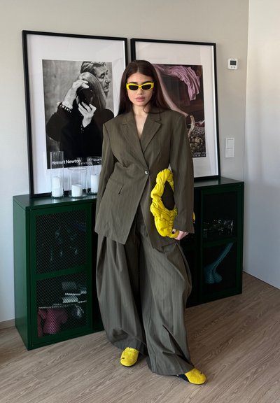 Mujer con traje de raya diplomática olivo de gran tamaño, gafas de sol amarillas, zapatos y bolso, de pie en una habitación con arte enmarcado y armario verde.