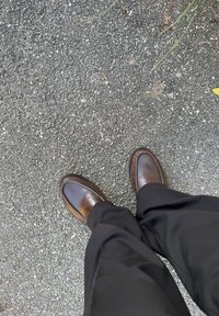 Mocassins en cuir marron avec couture mocassin, assortis à un pantalon noir, posés sur une surface asphaltée texturée.