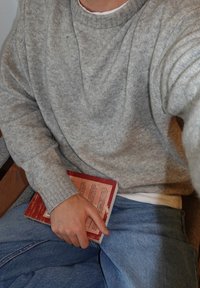 Pull en maille grise avec un col rond, associé à un jean denim bleu clair. Une main tient un livre rouge sur un fond neutre.