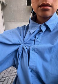 Camicia azzurro chiaro con dettaglio a nodo attorcigliato sul davanti, caratterizzata da diversi bottoni nello stesso tono e una texture di tessuto liscia.