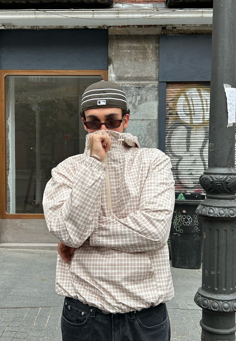 Coupe-vent à carreaux rose clair et beige avec des poignets élastiques et un col montant, superposé sur un t-shirt noir et un pantalon en jean.