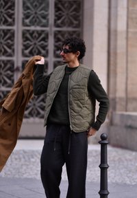 Olivovo zelená prešívaná vesta cez tmavý sveter, skombinovaná s čiernymi nohavicami. Drží hnedý kabát a stojí pri dekoratívnych kovových dverách.