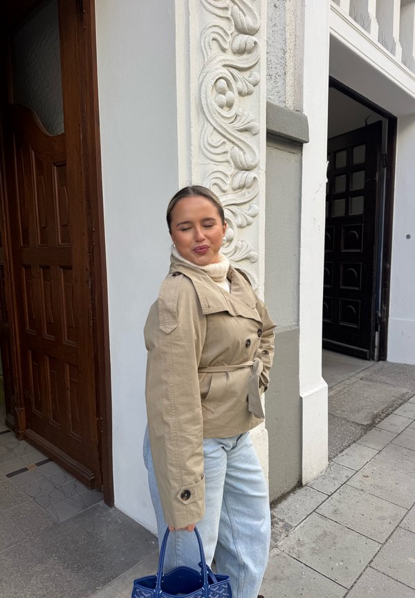 Jonge vrouw in een beige jas en lichte spijkerbroek houdt een blauwe handtas vast, staat naast een sierlijke witte pilaar en een gesloten houten deur, met gesloten ogen en een glimlach.