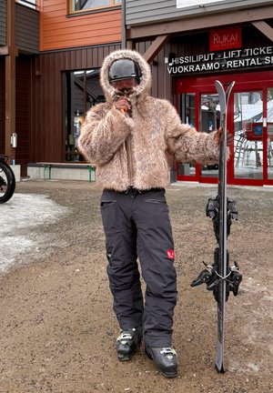 Persona con chaqueta peluda, gafas de esquí y botas sosteniendo esquís, parada frente a una oficina de alquiler y venta de billetes para remontes en una estación de esquí.