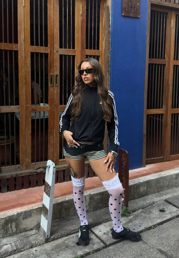 Femme aux longs cheveux portant une veste Adidas noire, un short rayé, des chaussettes hautes roses à pois, des chaussures noires et des lunettes de soleil, debout près de portes en bois.