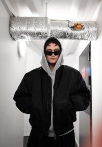 Juodas bomber tipo švarkas ant pilko džemperio su gobtuvu, derinamas su juodais saulės akiniais. Fone matomas metalinis oro kanalas ir uždarytos durys.