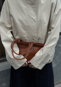 Kootud pruun nahast käekott kahe ümmarguse sangaga, hoitud beeži jope vastu. Kotil on ristkülikukujuline kuju ja tekstureeritud pind.