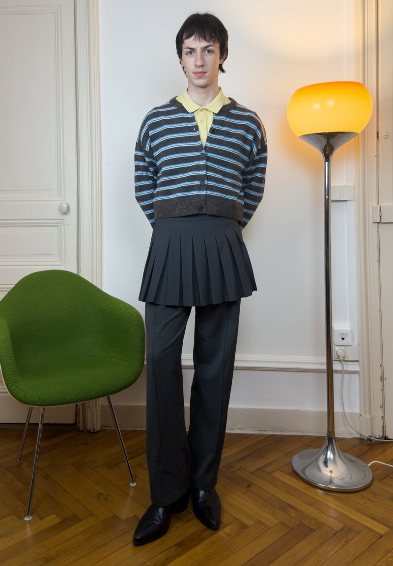 Young person standing indoors wearing a striped cardigan over a yellow shirt, black pleated skirt layered on pants, beside green chair and floor lamp.