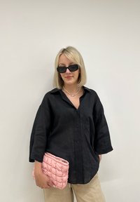Chemise noire à boutons avec coupe décontractée, pantalon en lin beige, pochette matelassée rose, et lunettes de soleil noires rectangulaires, sur fond blanc.