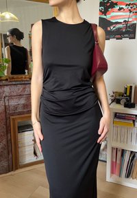 Femme portant une robe noire sans manches et un sac à bandoulière bordeaux, debout dans une pièce avec des étagères de livres, un miroir et des œuvres d'art murales colorées.