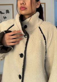 Krémová flísová bunda s čiernym lemovaním a zapínaním na gombíky. Dizajn s vysokým golierom; textúrovaný, mäkký materiál s kontrastným hnedým vnútrom.