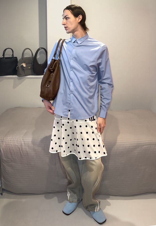 Personne portant une chemise bleu clair à boutons, jupe blanche à pois sur pantalon beige, chaussures bleues, tenant un sac à main marron sur l'épaule à l'intérieur.