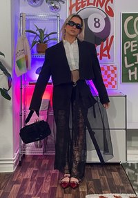 Crni skraćeni sako preko bijele košulje, upareno s prozirnim crnim hlačama. Dodaci uključuju crnu torbu i crvene balerinke sa zakovicama.