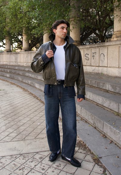 Joven con chaqueta de cuero color oliva, camiseta blanca, vaqueros oscuros y zapatos negros, de pie cerca de escalones de hormigón al aire libre con árboles al fondo.